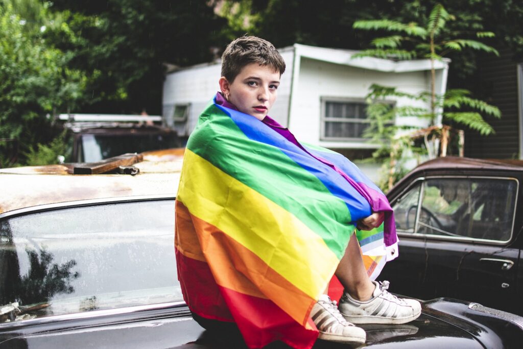 Une personne avec un drapeau arc-en-ciel assise sur le coffre d'une voiture, transformation homme-femme