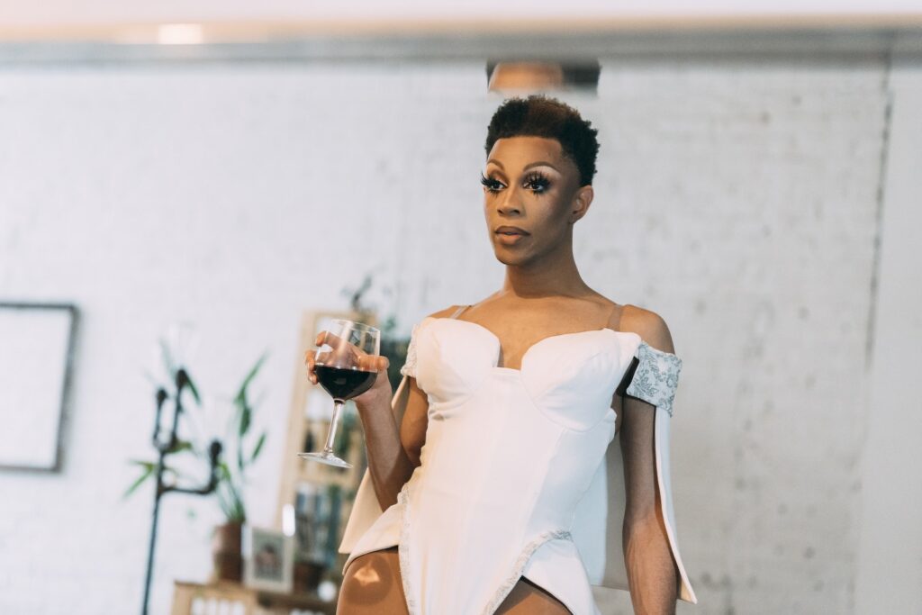 Une drag queen afro-américaine calme, vêtue d'une tenue élégante, tient un verre de vin rouge et se regarde dans un miroir dans une pièce lumineuse.