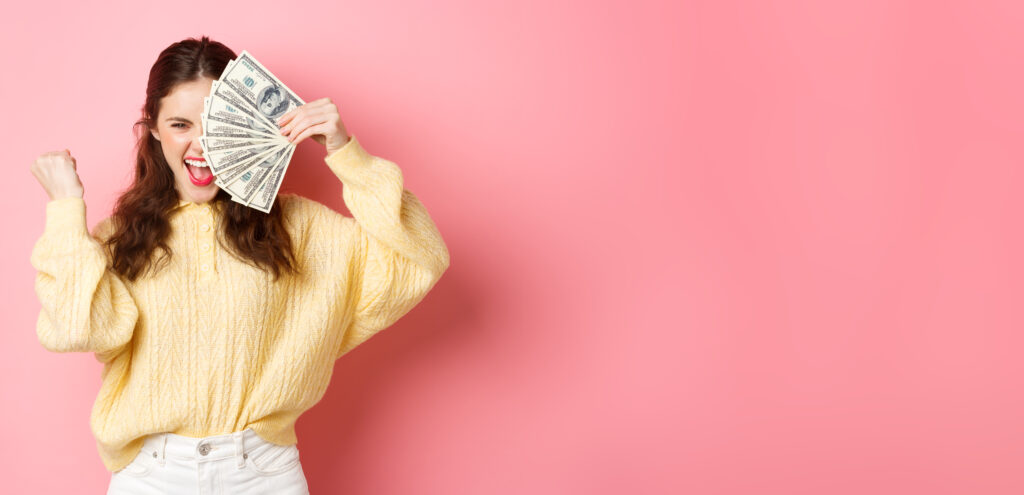 excited beautiful girl saying yes earn prize money holding dollar bills half face making fist pump gesture with satisfied expression standing against pink background