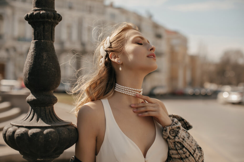 portrait beautiful blonde lady stylish white silk dress checkered jacket pearl necklace gently touching neck posing city square