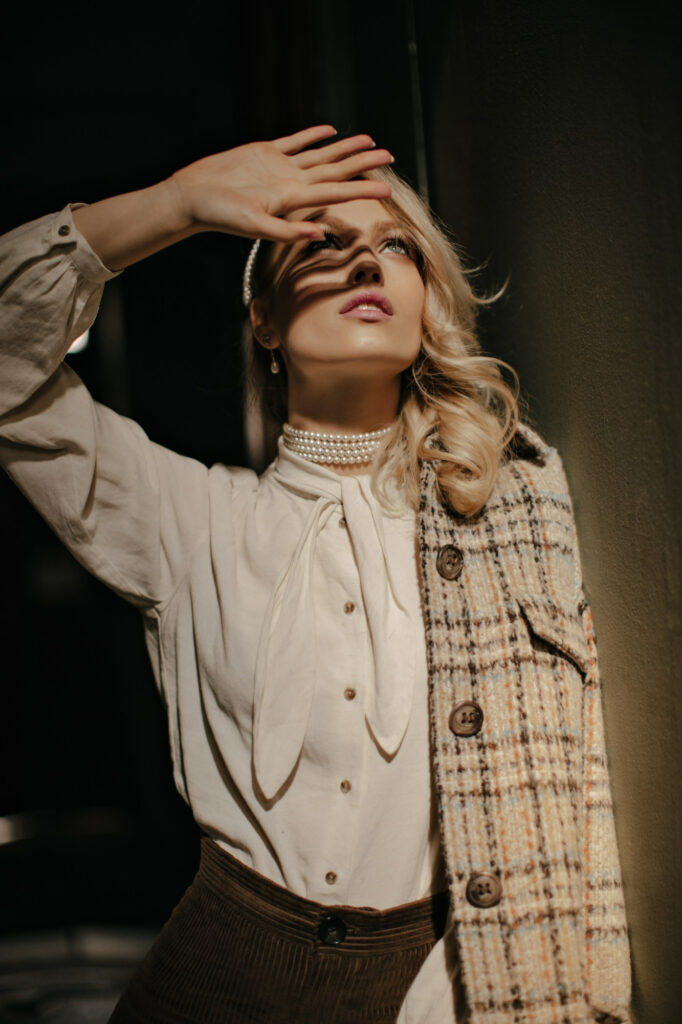 blonde curly woman tweed jacket white elegant blouse covers sun with hand charming girl looks up poses dark room