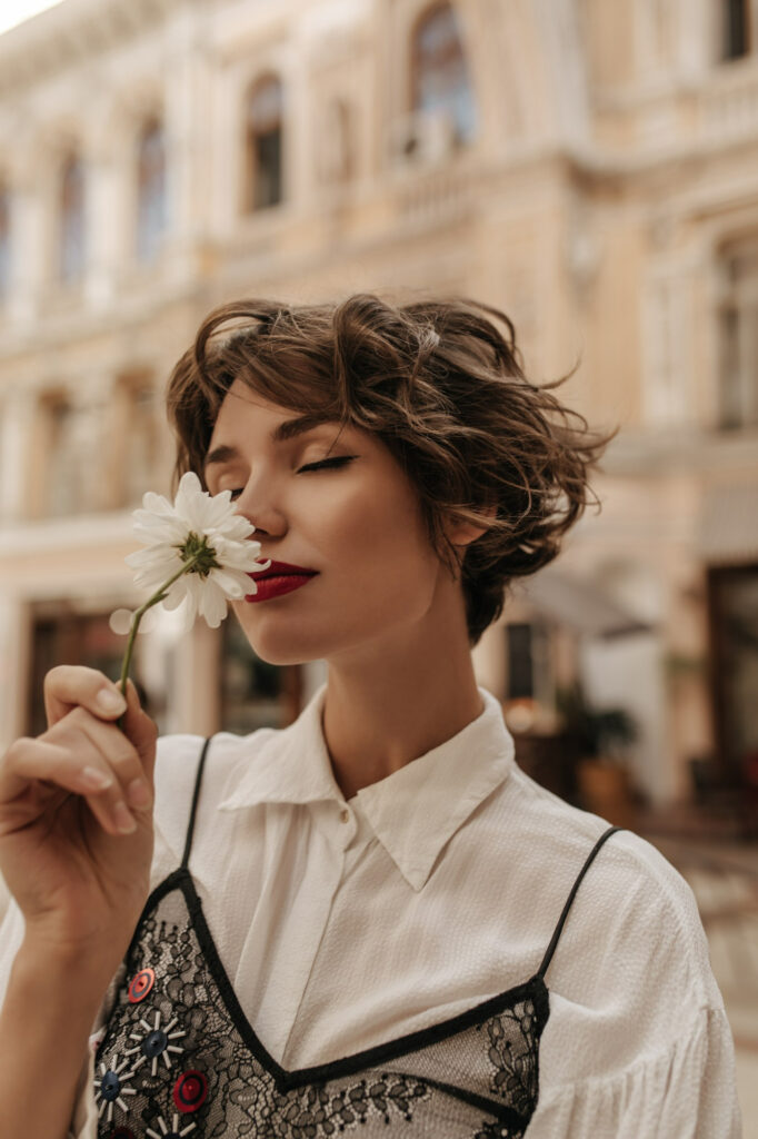 How Can I Make Myself Feel More Feminine 8 wavy haired woman light shirt with black lace sniffing flower city tender woman with red lips short hair poses street