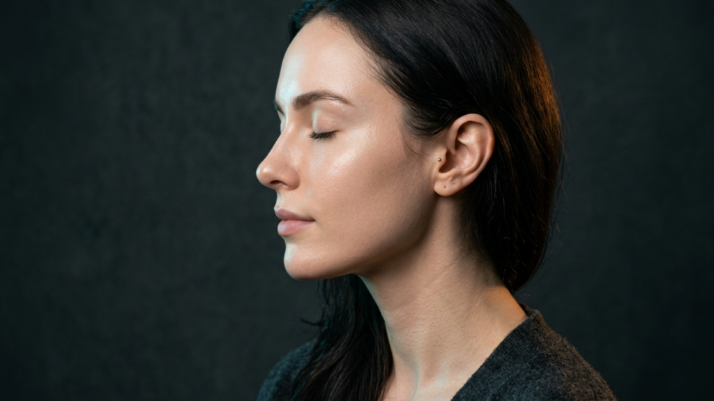 Un retrato hiperrealista en 8K de una mujer de perfil, capturado con la precisión de un objetivo fijo de 85 mm. La imagen presenta una iluminación de claroscuro magistral, con una dramática luz lateral que acentúa la elegante curvatura de su mandíbula y la textura suave y luminosa de su tez. Su piel muestra una luminosidad excepcional, resaltando los sutiles detalles de los poros y un brillo natural y saludable. Mantiene una expresión serena y meditativa, con los ojos suavemente cerrados, enfatizando un estado de ánimo tranquilo y contemplativo. La modelo viste una prenda de punto gris carbón oscuro con una textura sutil que contrasta con la suavidad de su piel. El fondo es minimalista, oscuro y apagado, lo que permite que el sofisticado juego de luces doradas en su cabello y sien se convierta en el punto focal, evocando una estética editorial íntima y de alta costura.